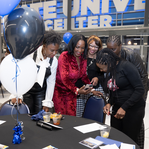 A group of five women, dressed formally, gather around a table looking at a smartphone during a Black Alumni Reception at Indiana State University. Blue and white balloons decorate the table.